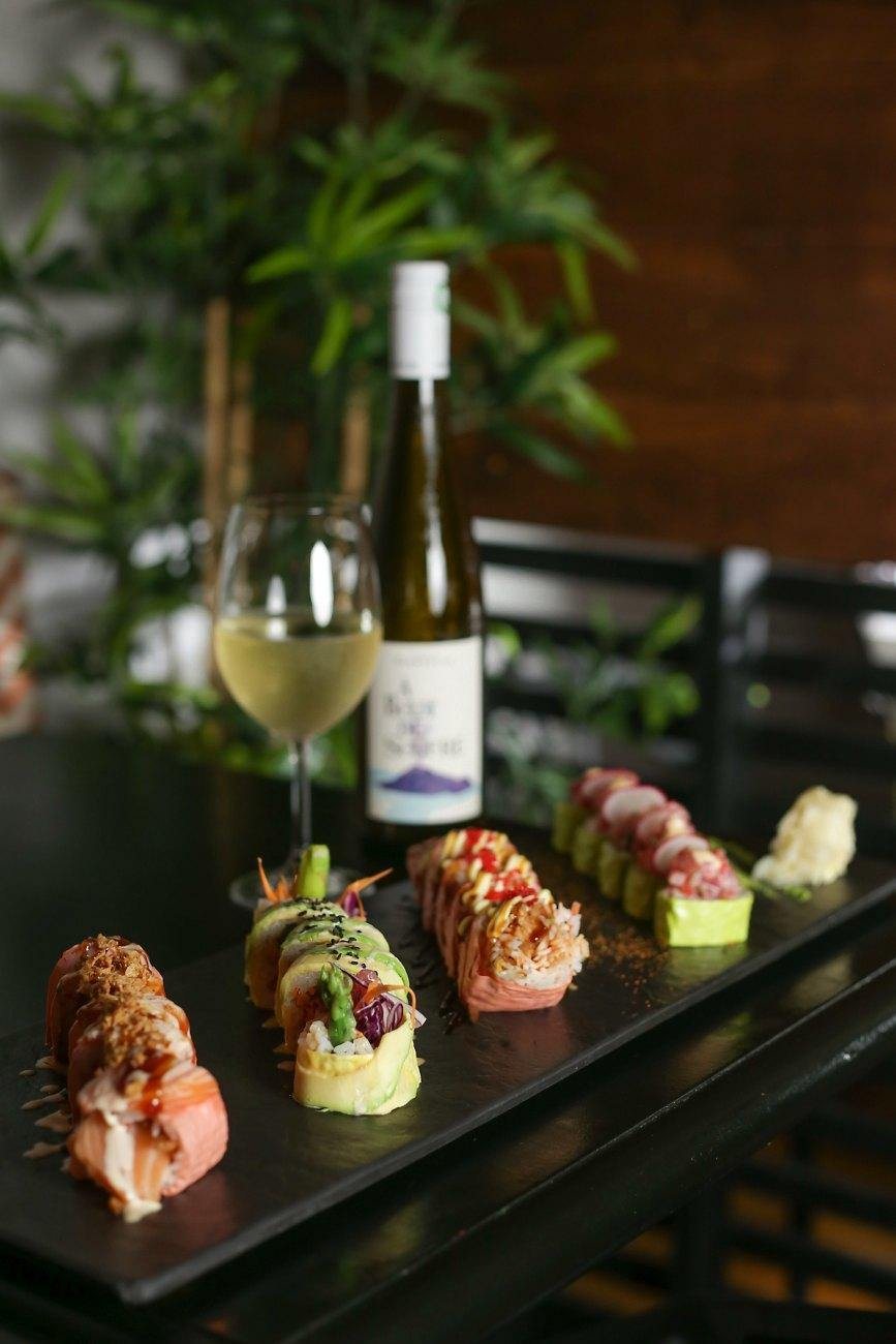 Makis lined up on a board beside a glass and a bottle of white wine